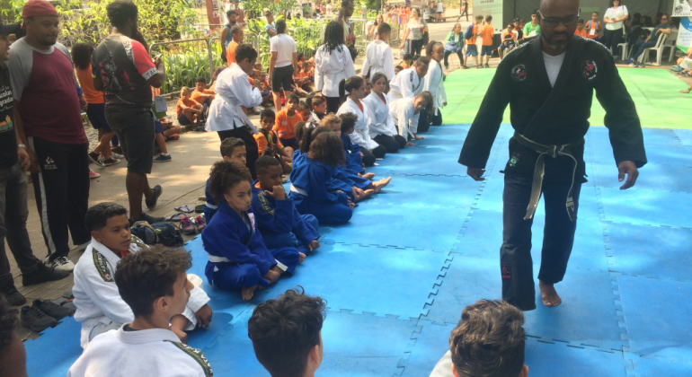 Na quinta-feira, dia 19/9, os estudantes da Rede Municipal mostraram a arte da luta e da dança na Praça da Estação e no Parque Municipal. Foto: Divulgação PBH Professor de judô, de pé, olha para mais de quinze alunos sentados, observados por mais gente, em local aberto, durante o dia, na Semana da Educação.
