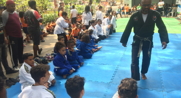 Professor de judô, de pé, olha para mais de quinze alunos sentados, observados por mais gente, em local aberto, durante o dia, na Semana da Educação. 