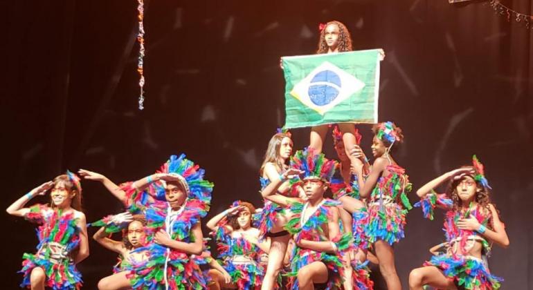 Dez crianças com fantasias de índio fazem apresentação em um palco e uma delas segura uma bandeira do Brasil