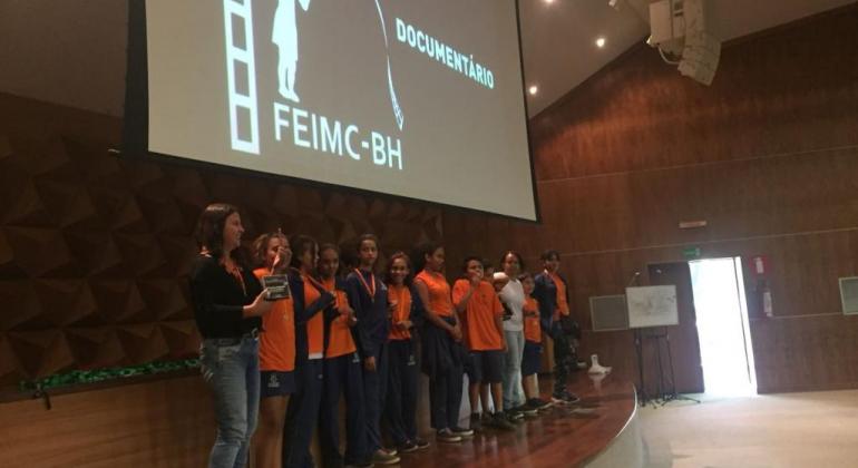 Imagem mostra uma mulher e cerca de 10 alunos em um palco, à frente de uma imagem projetada do FEIMC-BH 