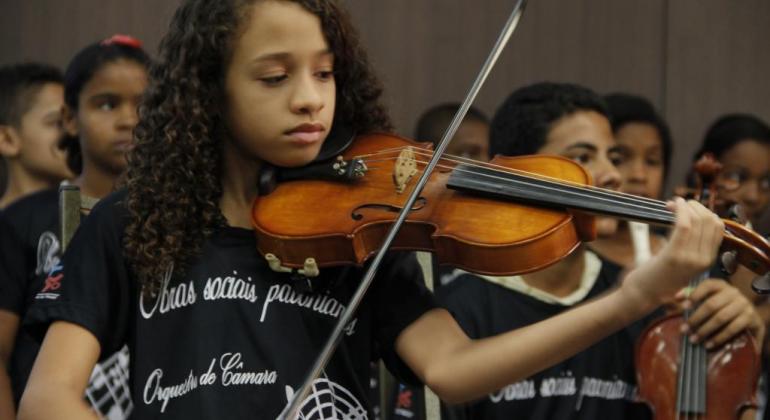 Foto mostra aluna tocando violino e cerca de seis alunos no palco tocando outros instrumentos 