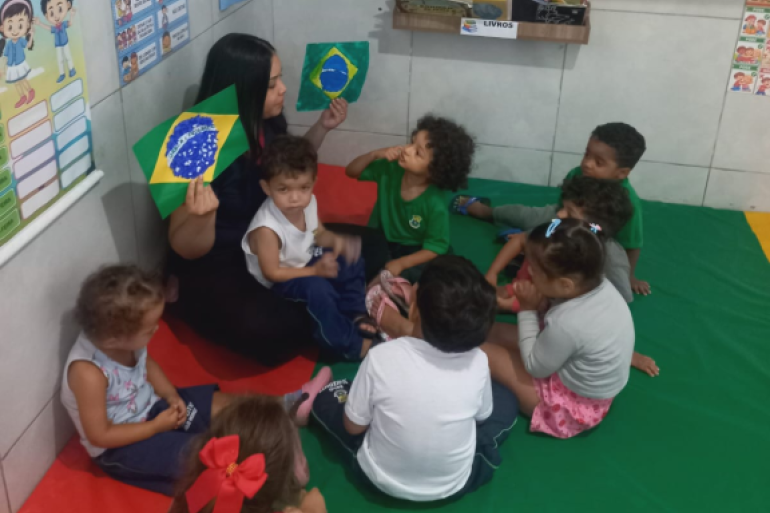 Crianças participam de atividade sobre a Copa do Mundo, explorando a bandeira do Brasil por meio de vivências lúdicas e interativas. #paratodosverem: Em sala de aula, professora sentada no chão mostra desenhos da bandeira do Brasil para crianças da Educação Infantil reunidas em roda sobre um tapete colorido. Os alunos observam a atividade, cercados por cartazes educativos e uma prateleira de livros.