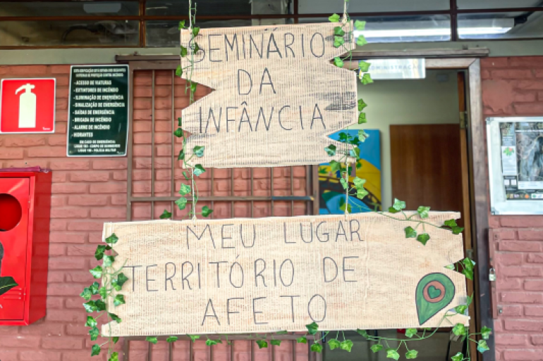 #paratodosverem: Porta de entrada da escola decorada com placas de madeira escritas “Seminário da infância” e “Meu lugar território de afeto”
