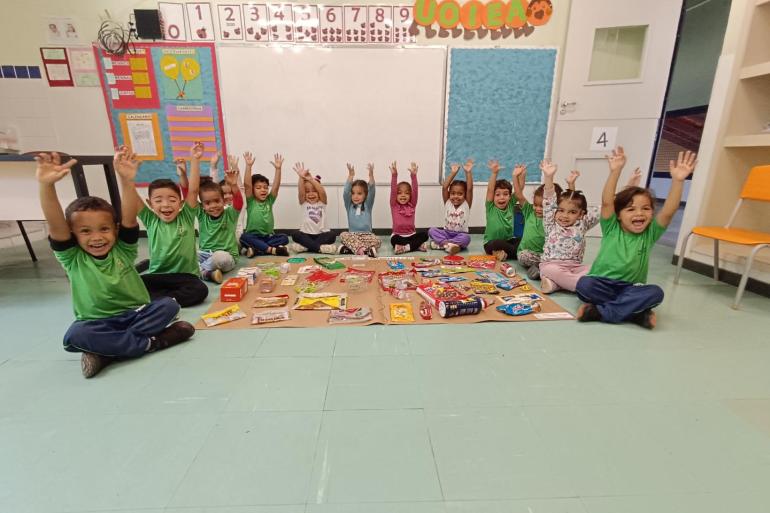 #paratodosverem: Em uma sala de aula, treze crianças pequenas estão sentadas no chão, em semicírculo, com os braços erguidos e sorrindo para a foto. À frente delas, há diversos produtos alimentícios, como biscoitos, salgadinhos e embalagens variadas, utilizados em uma atividade pedagógica. Ao fundo, há um quadro branco e painéis coloridos.