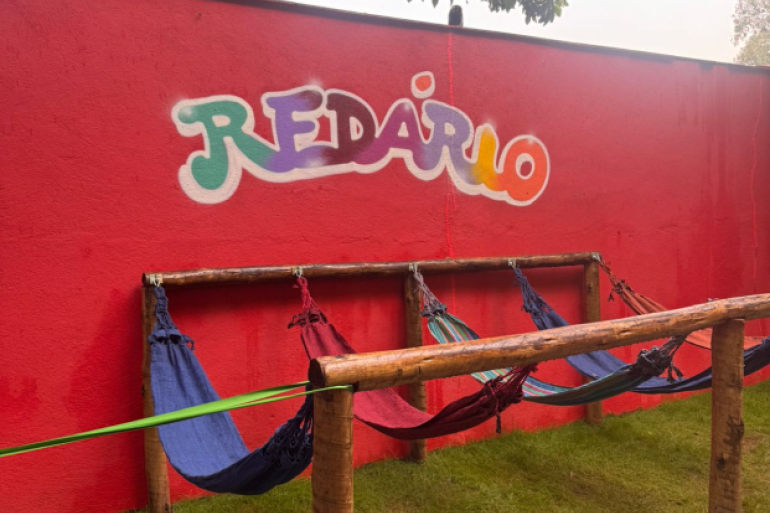 #paratodosverem: Área externa com uma parede vermelha onde está escrito “Redário” em letras coloridas. À frente, há uma estrutura de madeira com várias redes penduradas. O espaço é cercado por grama e árvores, criando um ambiente ao ar livre voltado para descanso e leitura.