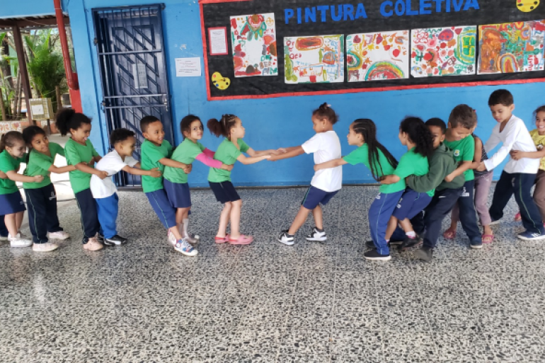 #paratodosverem: Um grupo de crianças, em fila, segura a cintura umas das outras e brinca em um pátio escolar. Elas usam uniformes verdes, azuis e brancos. Ao fundo, há um mural colorido com pinturas feitas pelas próprias crianças.