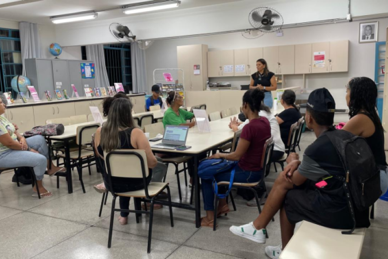 #paratodosverem: Em uma biblioteca escolar, um grupo de alunos da EJA está sentado ao redor de mesas organizadas em formato de reunião. Uma mulher está em pé à frente, falando e conduzindo a atividade. Sobre as mesas, há papéis e um notebook. Ao fundo, estantes com livros e materiais organizados indicam o espaço da biblioteca.