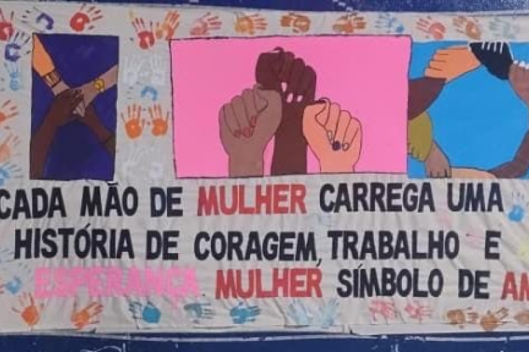 #paratodosverem: Painel colorido com ilustrações de mãos dadas e erguidas, representando união e diversidade. Ao centro, há a frase: “Cada mão de mulher carrega uma história de coragem, trabalho e esperança. Mulher símbolo de amor.” O fundo é decorado com várias marcas de mãos coloridas, sugerindo participação coletiva.