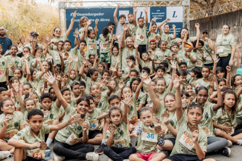 #paratodosverem: Crianças e adultos reunidos após uma corrida escolar. Todos usam camisetas verdes e muitos levantam as mãos e mostram medalhas, sorrindo para a câmera. Ao fundo, há um painel da escola.
