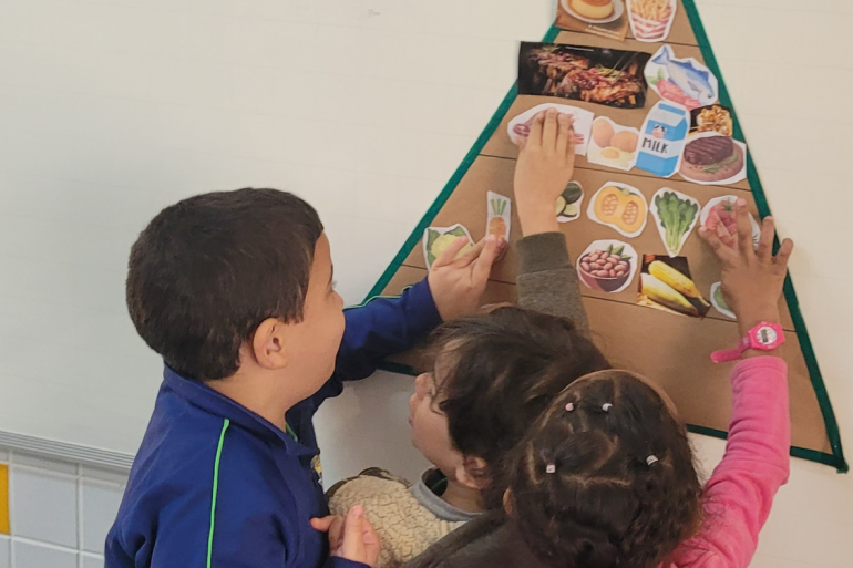 Crianças participam da construção da pirâmide alimentar, identificando os diferentes grupos de alimentos e aprendendo sobre escolhas mais saudáveis #paratodosverem: Três crianças participam de uma atividade em sala de aula, organizando figuras de alimentos em uma pirâmide alimentar fixada na parede. Enquanto colam as imagens, aprendem sobre os grupos alimentares e a importância de uma alimentação saudável de forma colaborativa e lúdica