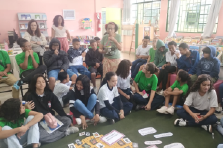 #paratodosverem: Alunos sentados em círculo no chão participam de uma atividade de leitura e conversa, com livros e cartões no centro da roda.