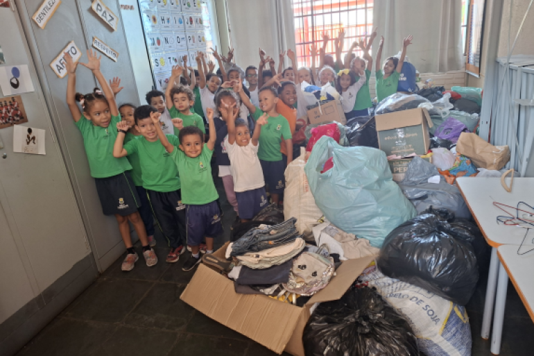 #paratodosverem: Crianças em sala de aula posam sorrindo ao lado de roupas, alimentos e brinquedos arrecadados para famílias atingidas pelas chuvas. 