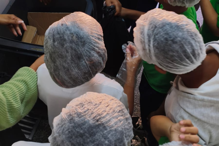 #paratodosverem: Grupo de estudantes reunidos ao redor de uma lixeira. As crianças usam toucas descartáveis e observam a atividade. A ação acontece na área da cozinha da escola e integra o projeto de conscientização sobre o desperdício de alimentos. 
