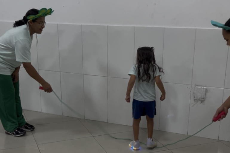 #paratodosverem: Em sala de azulejos brancos, duas adultas seguram uma corda no chão. À esquerda, mulher de calça verde usa tiara de folhas verdes. No centro, criança de costas com shorts azuis e tênis de luzes azuis acesas está sobre a corda. À direita, outra adulta usa a mesma tiara.