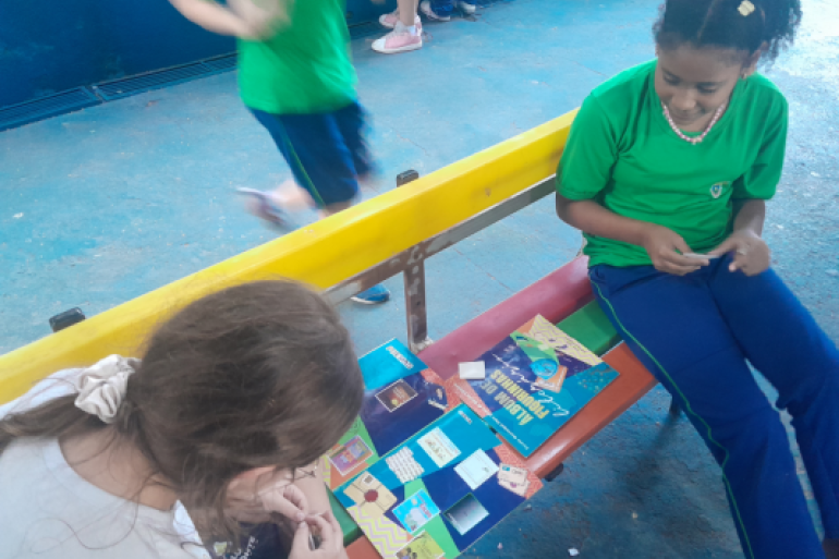 Além da leitura, os estudantes aprendem educação financeira #paratodosverem: Duas alunas sentadas em banco colorido com encosto amarelo organizam um "Álbum de Figurinhas Literárias" aberto entre elas. À direita, uma estudante de camiseta verde e calça azul manuseia figurinhas. À esquerda, em primeiro plano, outra estudante observa o álbum e cola um adesivo. O chão da área externa é azul. Ao fundo, outra criança em movimento passa correndo.