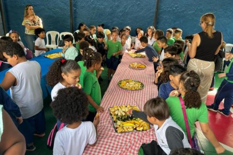 #paratodosverem: Crianças reunidas ao redor de mesas participam de uma atividade sobre alimentação saudável em ambiente escolar. Sobre toalhas xadrez vermelhas e brancas, há quatro bandejas com frutas variadas. Enquanto experimentam os alimentos, os alunos interagem entre si e são acompanhados por educadoras.