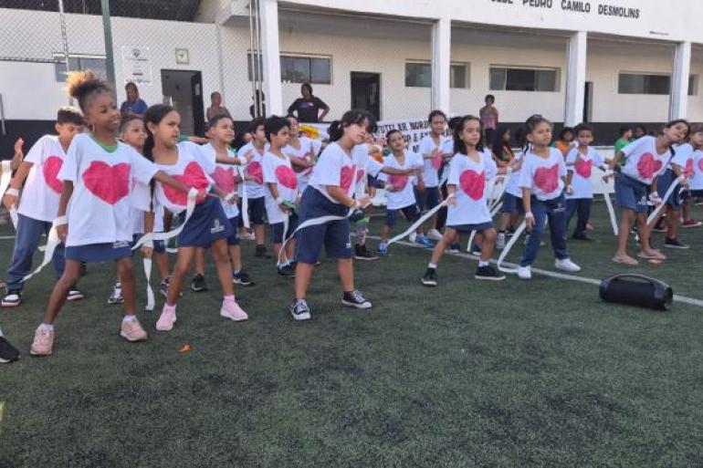 #paratodosverem: Grupo de crianças faz uma apresentação em campo de futebol. Elas usam camisetas brancas com desenhos de coração rosa e seguram fitas brancas nas mãos durante dança