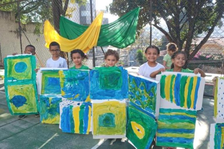#paratodosverem: Crianças exibem pinturas em tons de verde, amarelo e azul durante atividade ao ar livre. Os trabalhos são inspirados nas cores da bandeira do Brasil e estão organizados lado a lado. Ao fundo, tecidos verde e amarelo decoram o espaço.