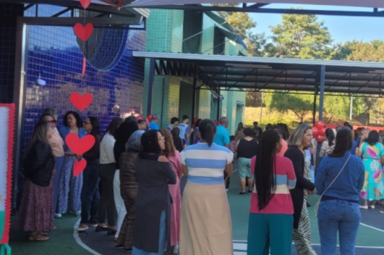 Avós participam de atividade especial na escola. #paratodosverem: Foto mostra um grupo de adultos reunidos na área externa da EMEI Belmonte, em um espaço coberto. As pessoas conversam e circulam próximas à quadra. O ambiente está decorado com corações vermelhos pendurados e balões. Ao fundo, aparecem paredes coloridas da escola, árvores e parte da cobertura metálica. Uma das participantes veste uma blusa listrada.