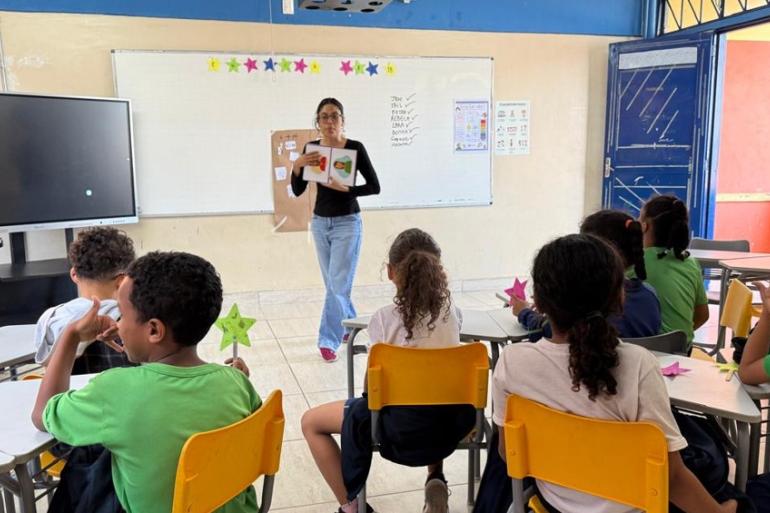 Estudantes da EMUG em aula do projeto de ensino bilíngue crianças sentadas em cadeiras de encosto amarelo, de costas para a imagem e de frente para a professora, que segura um material