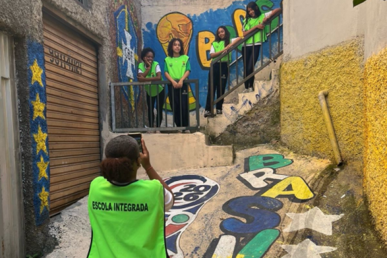 #PARATODOSVEREM: Quatro estudantes com coletes verdes posam em uma escadaria cercada por muros grafitados. Em primeiro plano, outra aluna fotografa o grupo com um celular. O local possui pinturas coloridas no chão e nas paredes, além de corrimãos e construções ao redor. 