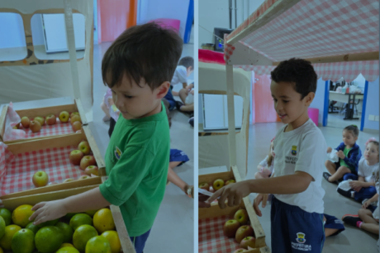 #PARATODOSVEREM: Duas crianças em um espaço montado com bancas de madeira decoradas com tecido xadrez. Os alunos observam e tocam frutas como laranjas, limões e maçãs dispostas nas prateleiras. Ao fundo, outras crianças aparecem sentadas no chão da sala acompanhando a atividade.