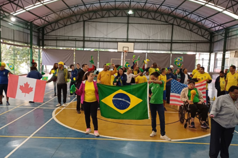#paratodosverem: grupo de pessoas reunido em uma quadra poliesportiva coberta celebrando com entusiasmo. Elas seguram bandeiras de diferentes países, como Brasil, Canadá, Estados Unidos e México, além de adereços verdes e amarelos