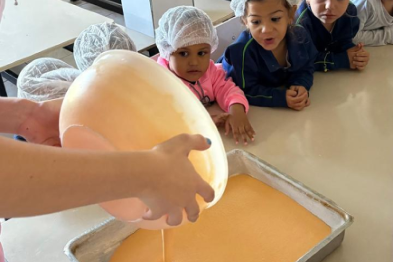 Crianças trabalham em equipe para produzir bolo de cenoura #paratodosverem: Em uma cantina, crianças pequenas usando toucas acompanham atentamente o preparo de um bolo apoiadas em mesa. Em primeiro plano, uma pessoa despeja a massa em uma forma.