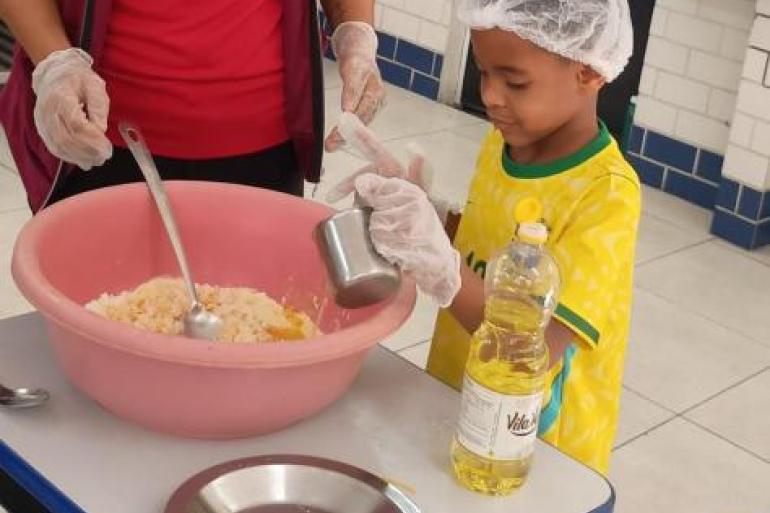 Alunos participam de atividade culinária na escola #paratodosverem: Menino usando touca e luvas descartáveis durante uma atividade culinária. Ao lado de uma educadora, ela segura um copo ao lado de uma mesa com uma tigela rosa contendo ingredientes e uma garrafa de óleo.