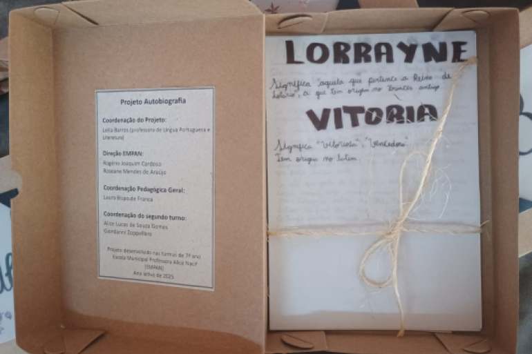 Histórias que cabem numa caixa, mas transbordam significado #paratodosverem: Uma caixa de papelão aberta com um livro artesanal amarrado com barbante, trazendo o nome “Lorrayne Vitória” na capa e informações sobre um projeto escolar de autobiografia.