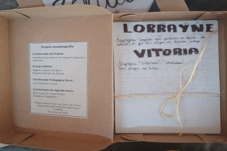 Histórias que cabem numa caixa, mas transbordam significado #paratodosverem: Uma caixa de papelão aberta com um livro artesanal amarrado com barbante, trazendo o nome “Lorrayne Vitória” na capa e informações sobre um projeto escolar de autobiografia