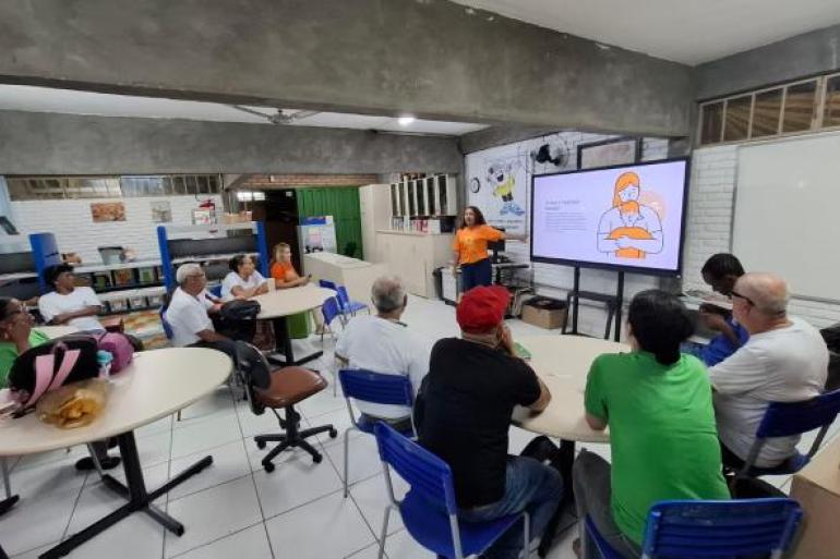 Atividade promove conscientização sobre violência infantil #paratodosverem: Em uma sala com mesas e cadeiras, homens e mulheres alunos da EJA estão sentados acompanhando uma palestra. Ao fundo, uma mulher de camiseta laranja, do Maio Laranja, apresenta conteúdos em uma tela digital sobre prevenção à violência e proteção de crianças e adolescentes. O ambiente possui estantes, quadro branco e materiais educativos.
