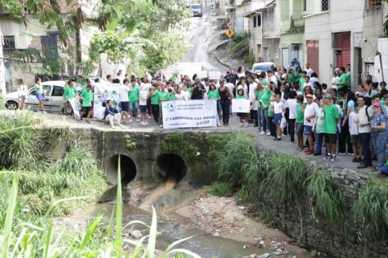 #paratodosverem: Grande grupo de estudantes e educadores reunidos em uma rua, ao lado de um córrego canalizado, durante uma caminhada. Os participantes seguram faixas e cartazes, com destaque para o banner “2ª Caminhada das Águas”, da Escola Municipal Rui da Costa Val, datado de 24-03-2026. Ao redor, há casas, vegetação e uma ponte com duas manilhas por onde passa a água.