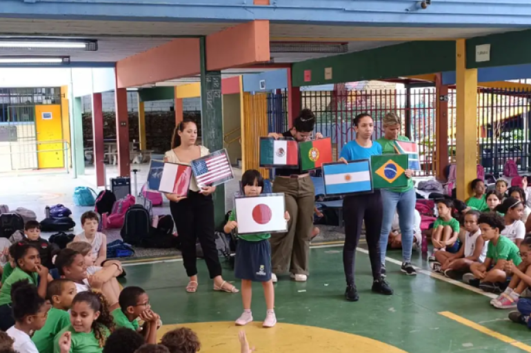 Alunos e educadores apresentam bandeiras no projeto "Volta ao Mundo". #PARATODOSVEREM: No pátio coberto da escola, dezenas de crianças estão sentadas no chão em semicírculo assistindo à atividade. Ao centro, uma aluna e quatro educadoras seguram cartazes com bandeiras de países como Japão, França, EUA, México, Portugal, Argentina e Brasil. O ambiente escolar é colorido, com colunas em tons de azul, amarelo e vermelho, além de mochilas organizadas ao fundo.