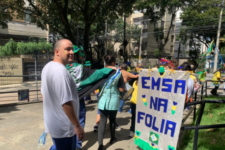 #paratodosverem: Um grupo de pessoas caminha em um desfile ao ar livre. Elas usam roupas coloridas e carregam um cartaz com a frase “EMSA na folia”, decorado com corações e a bandeira do Brasil. Ao fundo, aparecem prédios.