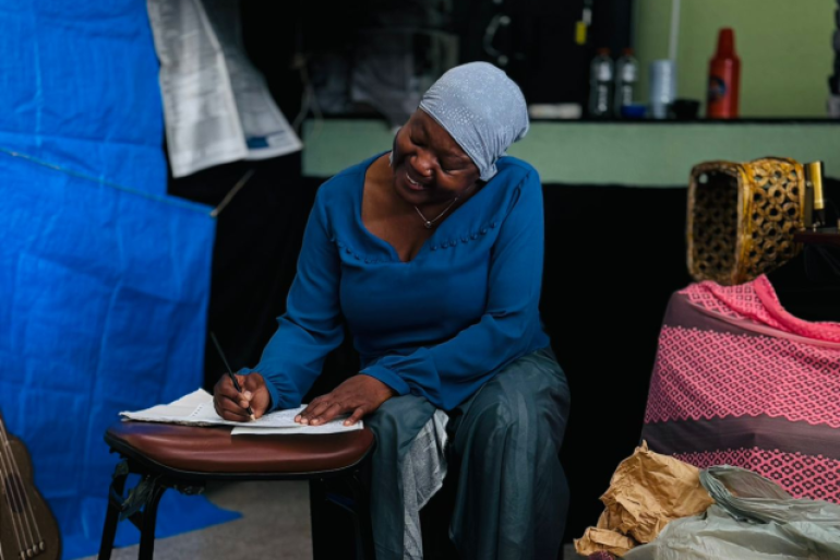 #paratodosverem: Mulher negra, sentada em um banco, usa um lenço na cabeça e roupa azul. Ela escreve concentrada em um caderno apoiado em um banquinho. O espaço ao redor tem objetos como sacolas, uma cadeira e um violão ao fundo, sugerindo um cenário de teatro que remete ao cotidiano retratado na obra.