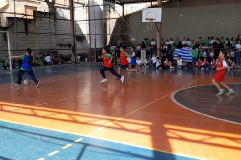 #paratodosverem: Em quadra poliesportiva bicolor (laranja e azul), acontece uma partida de futsal. Quatro estudantes em movimento vestem coletes vermelhos e azuis sobre seus uniformes. O estudante da esquerda, vestindo azul, conduz a bola. No fundo, há uma grade de proteção e uma arquibancada com muitos estudantes sentados assistindo. Uma cesta de basquete e uma trave de gol estão visíveis.