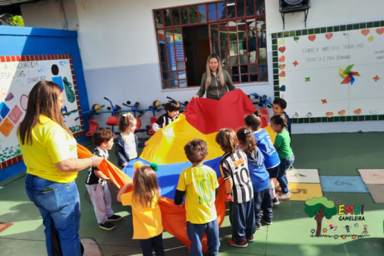 Diversão, movimento e aprendizado em equipe na Semana Esportiva da EMEI Gameleira #paratodosverem: Em uma área externa da escola, crianças participam de uma brincadeira com um grande pano com vários retalhos nas cores vermelho, amarelo, azul e laranja. Duas educadoras acompanham a atividade. Algumas crianças usam camisas de times de futebol. Ao fundo, há brinquedos infantis e murais decorados na parede.