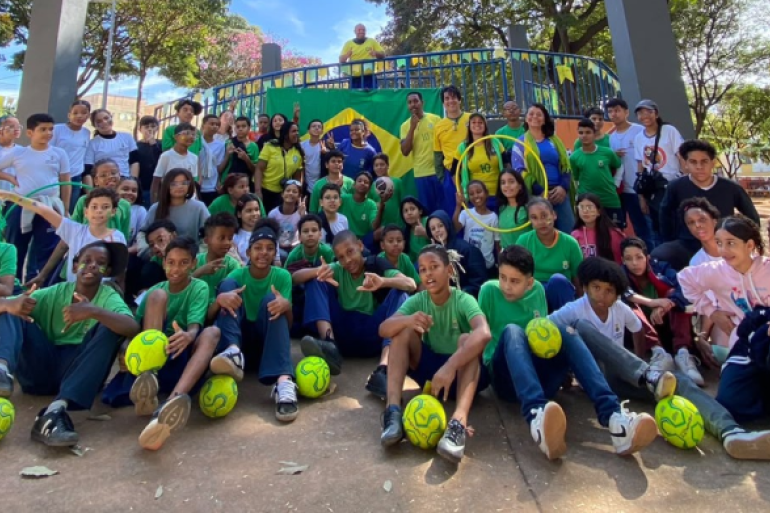 Comunidade escolar enfeita praça com as cores verde e amarelo #paratodosverem: Grupo de estudantes, professores e monitores do PEI posam para foto em uma praça. A maioria veste camisetas verdes, brancas e amarelas e alguns seguram bolas, bambolês. Uma bandeira do Brasil aparece ao fundo.