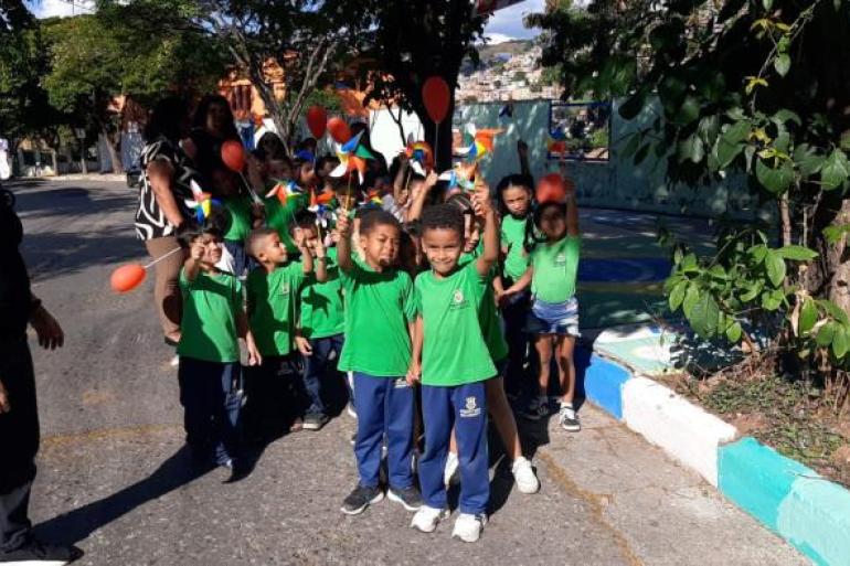 Crianças participam de caminhada educativa pelo bairro #paratodosverem: Grupo de crianças com uniformes escolares (camisas verdes e calças azuis) caminham acompanhados por adultos. Os alunos seguram cataventos coloridos e balões vermelhos, enquanto olham para a câmera.