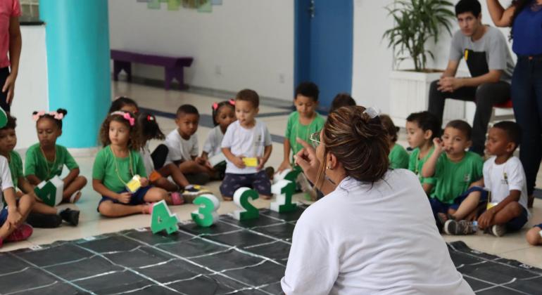  imagem mostra um grupo de crianças vestindo camisetas verdes e brancas, participando de uma atividade educacional em um ambiente escolar.