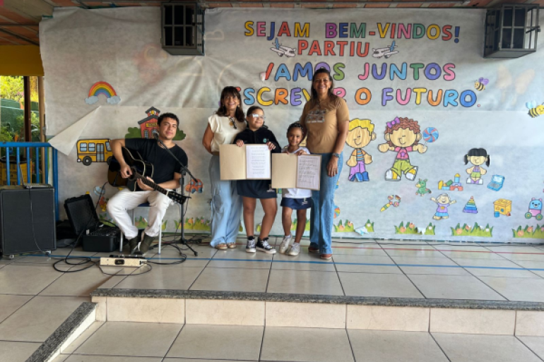 #paratodosverem: A foto mostra duas crianças ao centro segurando um cartaz aberto, acompanhadas por duas mulheres adultas ao lado delas. À esquerda, um homem toca violão sentado. Todos estão em frente a um painel com a frase “Sejam bem-vindos! Vamos juntos escrever o futuro.”