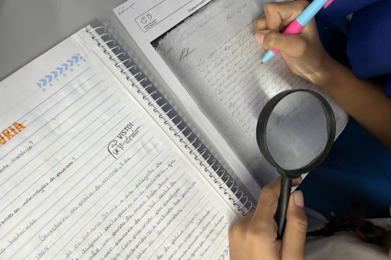 Entre o passado e o presente, estudantes investigam a história com atenção aos detalhes. #paratodosverem: Imagem vista de cima mostra as mãos de um estudante escrevendo em um caderno enquanto segura uma lupa sobre um documento antigo impresso. Ao lado, há outro caderno com anotações feitas à mão. A cena sugere uma atividade escolar de análise de fontes históricas.