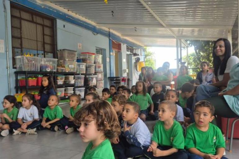 #paratodosverem: Em espaço coberto da escola, crianças pequenas uniformizadas se sentam no chão e observam atentamente uma atividade fora do enquadramento. Elas vestem uniformes nas cores verde, branco e azul. Educadoras e alguns responsáveis acompanham o momento também sentados.