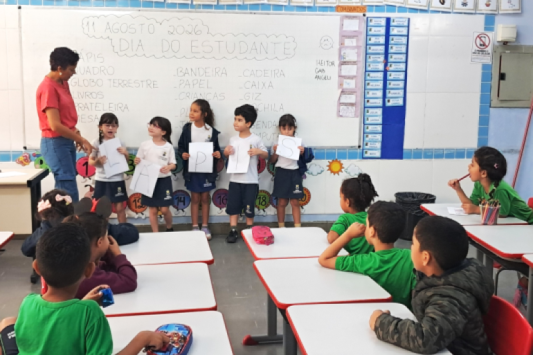 Atividade incluiu a leitura coletiva das letras. #paratodosverem: Em uma sala de aula, cinco alunos de uniforme estão alinhados em frente ao quadro branco segurando papéis com as letras L, Á, P, I, S, orientados por uma professora de blusa vermelha. No quadro está escrito "11 AGOSTO 2026 DIA DO ESTUDANTE". Em primeiro plano, outros alunos sentados em carteiras brancas com bordas vermelhas observam a apresentação. Acima do quadro, há um alfabeto.