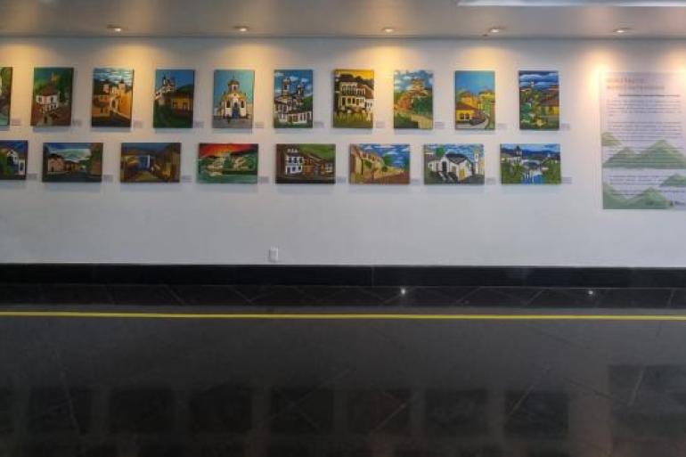 Exposição retrata lugares e paisagens históricas de Ouro Preto #paratodosverem: Exposição com 18 pinturas coloridas de paisagens e construções históricas expostas em uma parede branca da galeria. As obras retratam cenas de Ouro Preto, com igrejas, ruas e casas em diferentes estilos e cores. Ao lado, há um painel informativo com o título “Ouro Preto: Nosso Patrimônio”.