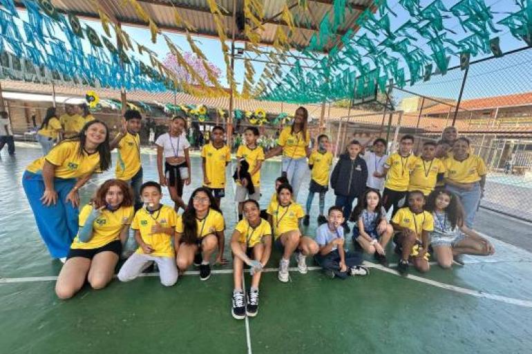 Espírito esportivo e jogo limpo unem estudantes de Emef #paratodosverem: Em uma quadra coberta e decorada com bandeirinhas do Brasil e das cores verde, amarelo e azul, estudantes e educadores posam para fotografia em grupo. A maioria veste camisetas amarelas inspiradas no uniforme da Seleção Brasileira e usa medalhas no pescoço. Algumas crianças estão sentadas à frente, enquanto outras permanecem em pé ao lado dos professores.