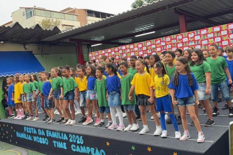 #paratodosverem: Um grupo com várias crianças se apresenta em um palco preto montado no pátio de uma escola. Elas vestem camisas verdes, azuis ou amarelas e shorts jeans. À frente do palco, está escrito "FESTA DA FAMÍLIA 2026 FAMÍLIA: NOSSO TIME CAMPEÃO". Ao fundo, há um painel decorado com corações vermelhos e brancos.