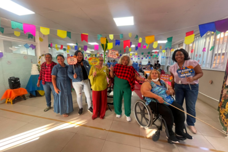 #paratodosverem: Sete mulheres posam sorridentes sob bandeirinhas coloridas de Festa Junina em um salão iluminado. Elas vestem trajes casuais e xadrez, segurando plaquinhas divertidas com frases como "MODO ARRAIÁ ATIVADO". Entre elas, uma mulher está em uma cadeira de rodas. Ao fundo, há mesas compridas organizadas para a celebração e uma caixa de som sobre uma pequena mesa laranja à esquerda.