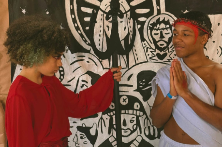 A releitura do clássico da literatura brasileira fez sucesso #paratodosverem: Dois jovens atuam em uma cena teatral. À esquerda, um jovem de cabelos cacheados e blusa vermelha interpreta o Diabo, segurando um tridente preto e olhando fixamente para o outro. À direita, um jovem de túnica branca, coroado com espinhos e mãos postas em oração, representa Jesus. Ao fundo, um painel ilustrado em estilo cordel em preto e branco exibe figuras sacras e nordestinas.